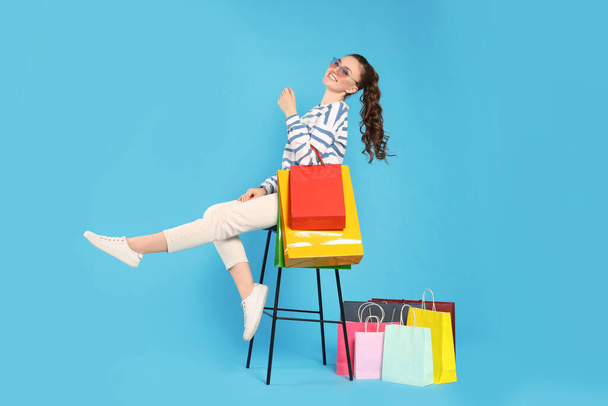 Happy woman in stylish sunglasses holding colorful shopping bags on stool against light blue background - Foto, imagen