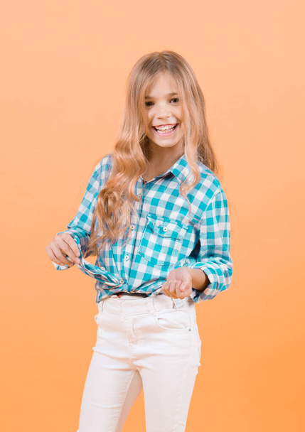 Girl smile in blue plaid shirt and white pants. Happy child with blond long hair on orange background. Kid fashion, beauty, style, look concept - Photo, Image