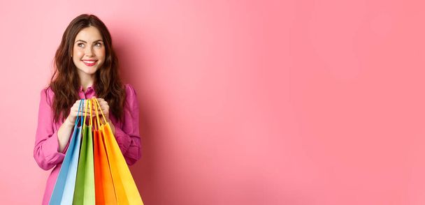 Fröhliche junge Frau denkt daran, etwas zu kaufen, hält mit verträumtem Lächeln Einkaufstüten in der Hand, kauft Personal, steht vor rosa Hintergrund. - Foto, Bild