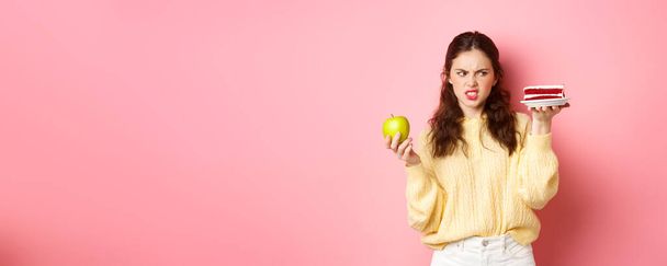 Gesundes Mädchen auf Diät schaut angewidert auf ein Stück Kuchen, hält grüne Applikation in der Hand, grimmig unzufrieden, steht vor rosa Hintergrund. - Foto, Bild