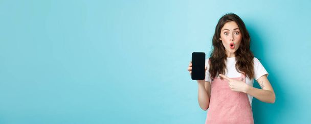 Etkilenmiş genç modern kadın akıllı telefon ekranını işaret ediyor, hayret verici ve hayret verici bir şekilde kameraya bakıyor, harika bir uygulama gösteriyor, yeni bir online mağaza, mavi arka planda duruyor.. - Fotoğraf, Görsel