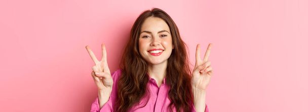 Lähikuva positiivisesta ruskeaverikkö naismallista, joka osoittaa rauhanmerkkejä, hymyilee ja pitää hauskaa, vain hyviä viboja, seisoo vaaleanpunaisen taustan päällä. - Valokuva, kuva