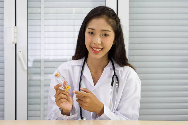 Beyaz önlüklü ve steteskoplu genç Asyalı doktor, hastaya kablosuz iletişim yoluyla sunulan tahta kapaklı cam hap kutusu taşıyor. Sağlık ve Tıp Konsepti. - Fotoğraf, Görsel
