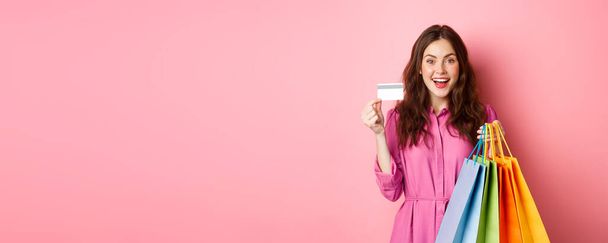 Portré izgatott boldog shopaholic, nő kezében bevásárlószatyrok és bemutató műanyag hitelkártya, mosolygós csodálkozva, áll szemben rózsaszín háttér. - Fotó, kép