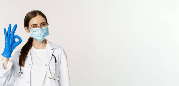 女性医師の肖像画を閉じます,コロナウイルスから顔のマスクの医師,承認で大丈夫な兆候を示します,推薦,賞賛と笑顔良いように,白い背景の上に立って. - 写真・画像
