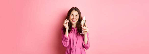 Concepto de celebración y festivos. Elegante mujer moderna en vestido de moda, sosteniendo la copa de champán en la fiesta, sonriendo complacido, de pie sobre el fondo rosa. Copiar espacio - Foto, Imagen
