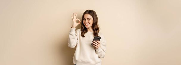 Beautiful woman holding cellphone, mobile phone and okay sign, recommending application, shopping app, standing over beige background. - Fotoğraf, Görsel