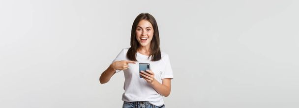 Excited and pleased brunette girl pointing finger at smartphone screen. - Фото, изображение
