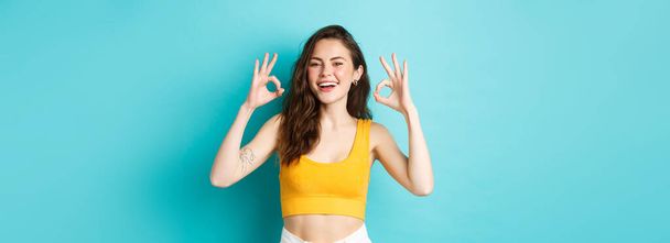 Young carefree woman say yes and nod in approval, showing okay gestures to praise good choice, show perfect body in summer outfit, ready for vacation, standing over blue background. - Photo, Image