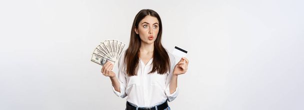 Excited woman holding credit card and money, looking amazed at cash, standing against white background. Copy space - Foto, Bild