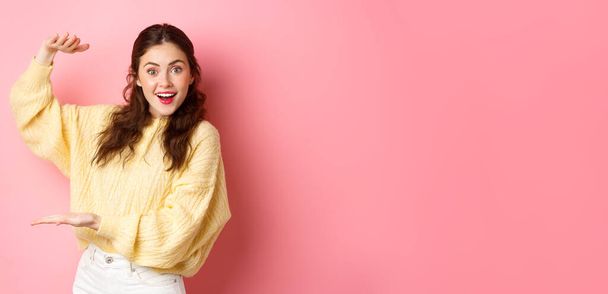 Jonge vrolijke vrouw staart naar camera, toont groot object, houdt uw logo of product met haar handen opzij op kopieerruimte, staande tegen roze achtergrond. - Foto, afbeelding