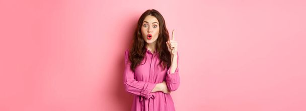 Young stylish feminine girl saying suggestion, have solution, got an idea, raising finger up in eureka sign, standing over pink background. - 写真・画像