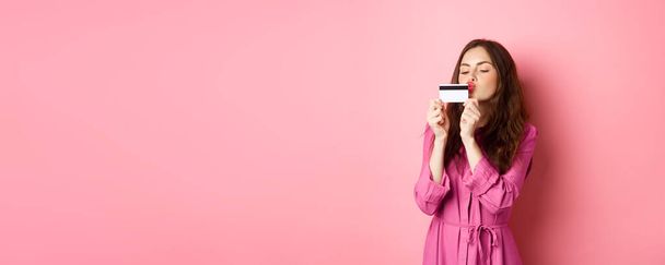 Image of young beautiful woman kissing her credit card, going on shopping, wasting money in stores, standing against pink background. - Foto, afbeelding