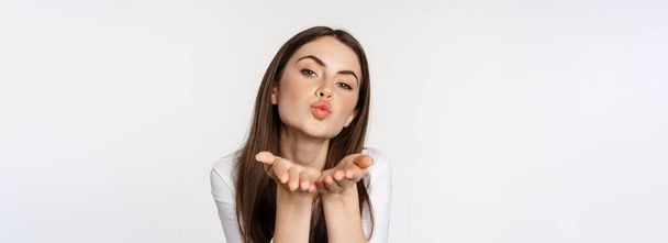 Feminine woman sending air kiss at camera, coquettish flirty pose, kissing, standing over white background. Copy space - Zdjęcie, obraz