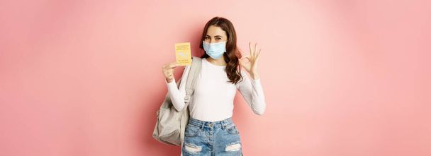 Młoda kobieta w masce medycznej, wykazująca się dobrym znakiem i zaświadczeniem o szczepieniu przeciw wiremii, podróżująca podczas pandemii, wyjeżdżająca na wakacje, stojąca na różowym tle. - Zdjęcie, obraz
