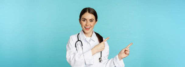 Retrato del trabajador médico sonriente, doctora en bata blanca con estetoscopio, señalando con los dedos a la izquierda, mostrando publicidad clínica médica, fondo turquesa. - Foto, Imagen