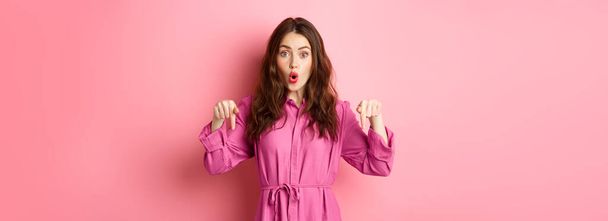 Stylish caucasian woman saying wow, pointing fingers down at impressive banner news, showing advertisement, staring amazed, standing against pink background. - Фото, зображення