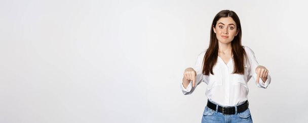 Doubtful cute girl shrugging unsure, smiling and pointing fingers down, standing over white background. - Photo, image