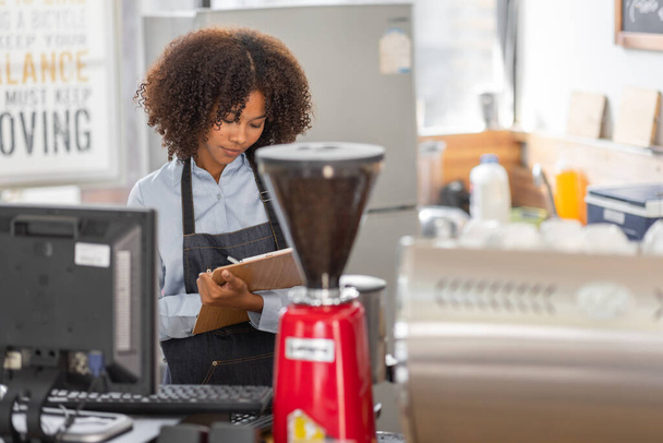 Vrouwelijke Afrikaanse coffeeshop kleine ondernemer draagt schort staan in de voorkant van teller uitvoeren van voorraadcontrole. afro haar werknemer Barista ondernemer. - Foto, afbeelding