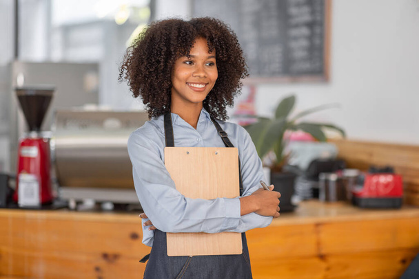 女性アフリカコーヒーショップカウンター前に立ってエプロンを身に着けている中小企業のオーナーは、株式チェックを実行します。アフロヘア従業員バリスタ起業家. - 写真・画像