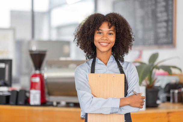 Female African coffee shop small business owner wearing apron standing in front of counter performing stock check. afro hair employee Barista entrepreneur. - Photo, Image
