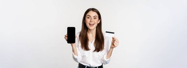 Glückliche Frau zeigt Kreditkarte und Smartphone-Bildschirm, Konzept des Online-Shoppings, Kauf in App, steht vor weißem Hintergrund. - Foto, Bild
