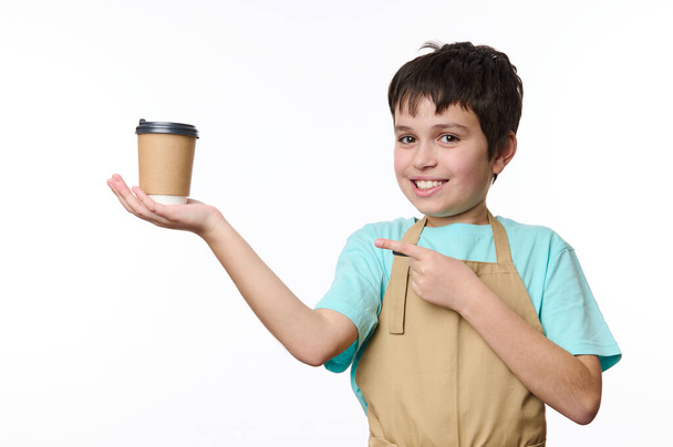 Entzückender Teenager, Barmann Barista Barkeeper mit beiger Kochschürze, der einen Kaffee zum Mitnehmen in Pappbecher verkauft. Heißgetränk to go, isoliert auf weißem Hintergrund. Kopierraum - Foto, Bild