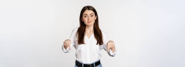 Doubtful cute girl shrugging unsure, smiling and pointing fingers down, standing over white background. - Photo, Image
