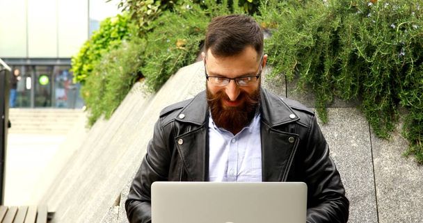 Ein bärtiger junger Mann arbeitet draußen am Laptop. Schöner Mann tippt auf Laptop-Tastatur auf der Straße. - Foto, Bild
