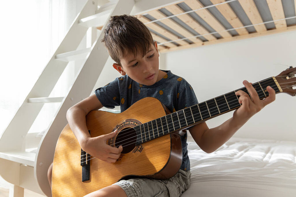 アコースティックギターを学ぶ少年。少年は自分の部屋でアコースティックギターを練習している - 写真・画像