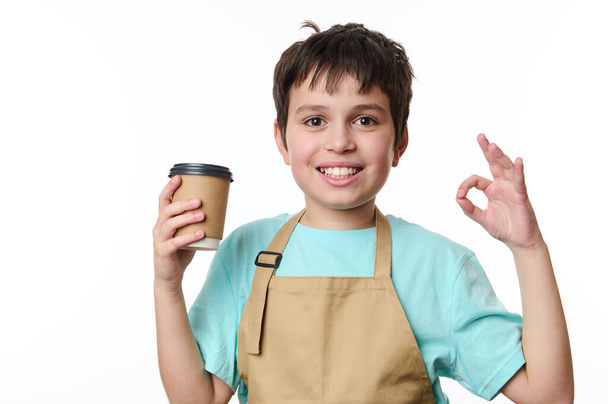 Ritratto isolato su sfondo bianco di un felice ragazzo barista adolescente che mostra segno OK, vendendo una bevanda da asporto caffè in tazza di carta ecologica. Bevanda calda da asporto. Copia lo spazio per la tua pubblicità - Foto, immagini