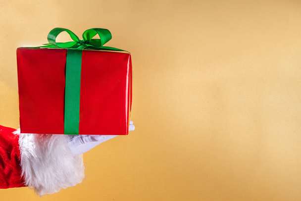 Boldog karácsonyi üdvözlőlapot, ajándékokat és értékesítési promóciós banner háttér. Mikulás kezek nagy piros ajándék doboz ünnepi zöld szalag, több mint arany háttér - Fotó, kép