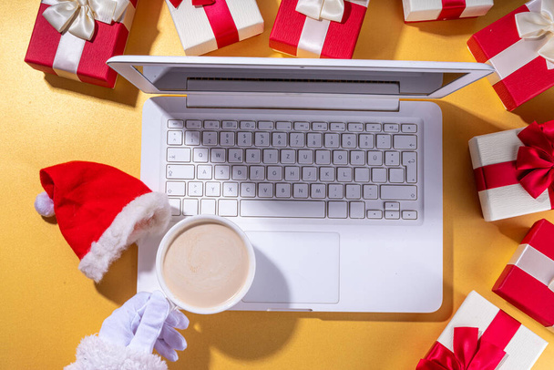 Vente de Noël, préparation pour les vacances concept. Les mains du Père Noël avec ordinateur portable, tasse de latte au chocolat chaud, boîtes-cadeaux, couché plat sur fond doré. Faire une liste de souhaits, préparer des cadeaux pour Noël. - Photo, image