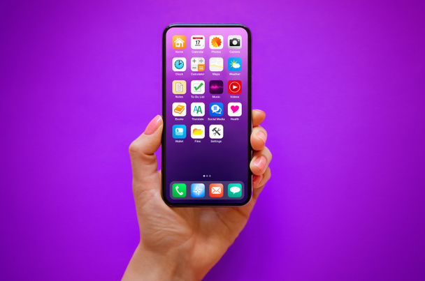 Mobilní telefon v ruce na fialovém pozadí se vzorkem ikony domácí obrazovky na obrazovce - Fotografie, Obrázek