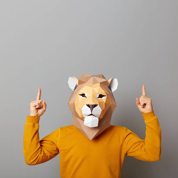 Image of man with lion mask with wearing casual style orange jumper, indicating upwards, showing copy space for advertisement, posing isolated over gray background. - Fotó, kép