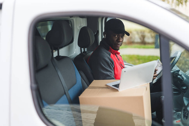 Beyaz minibüsün içinde oturan genç siyahi teslimatçı kağıtları dolduruyor ve laptopla pencereden kameraya bakıyor. Yatay çekim. Yüksek kalite fotoğraf - Fotoğraf, Görsel