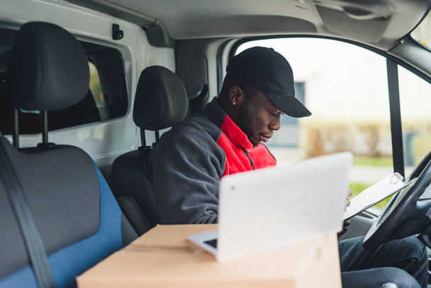 Young adult black delivery man sitting in driver seat of van looking through documents with open laptop computer on the side. Horizontal shot. High quality photo - Фото, зображення
