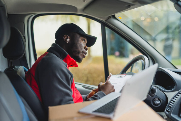 Young adult black delivery man sitting in driver seat of van holding documents and looking out the front window taking break from work. Horizontal shot. High quality photo - Φωτογραφία, εικόνα