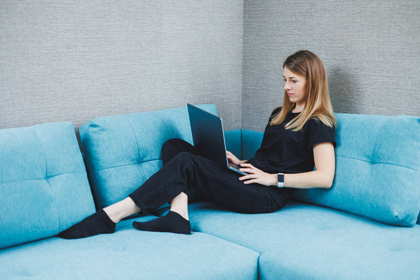 Una joven está sentada en un sofá y trabajando en un portátil. Trabajo remoto. Trabajar en casa en línea - Foto, Imagen