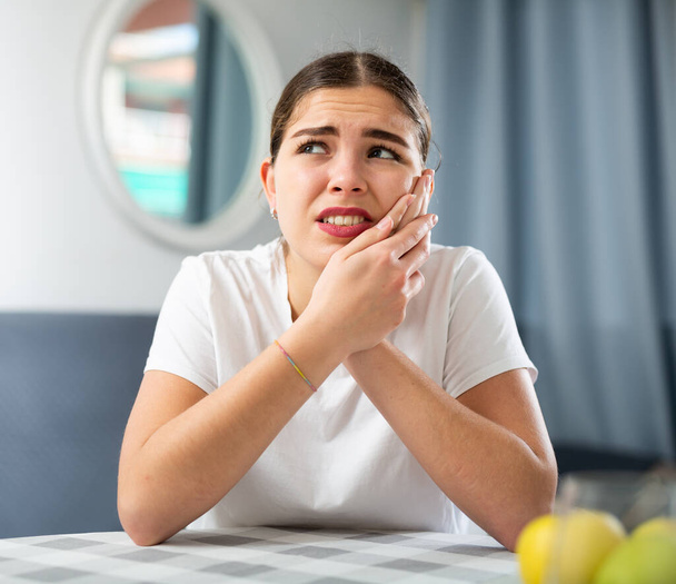 Portrait of a young european girl suffering from toothache and holding her hands on her cheek, sitting at a table in an ..apartment - Фото, изображение