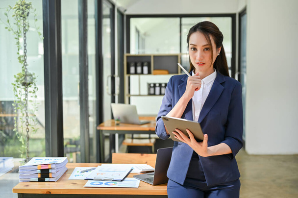 Urocza i atrakcyjna milenijna azjatycka bizneswoman lub szefowa w formalnym garniturze biznesowym trzymająca cyfrowy tablet, patrząca w kamerę, stojąca w swoim biurze. - Zdjęcie, obraz