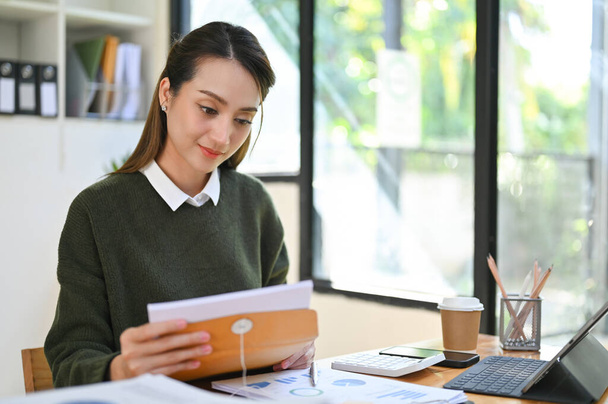 Güzel Asyalı iş kadını ya da kadın asistan okumak için mektubu ya da belgeyi açıyor, masasında çalışıyor.. - Fotoğraf, Görsel
