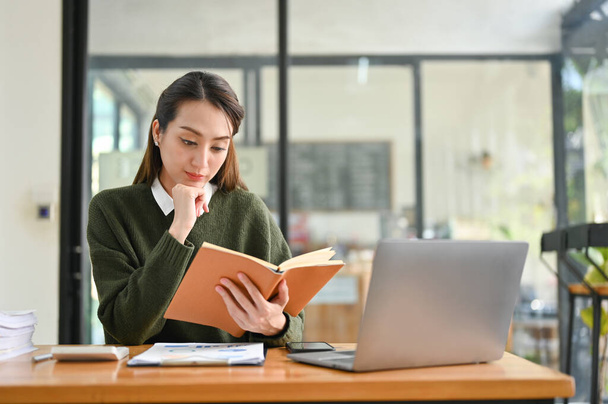 Determined and professional millennial Asian businesswoman or female financial worker reading a book, researching information in a textbook, working at her desk. - 写真・画像