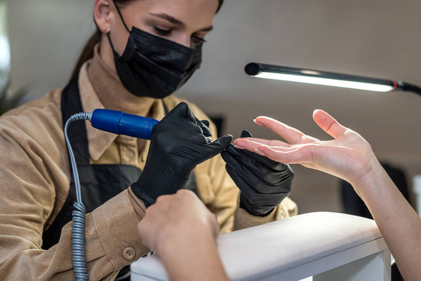 the master qualified on nails in prestigious salon in special gloves carries out sawing of a nail plate to the client. Manicure concept during a pandemic - Фото, зображення