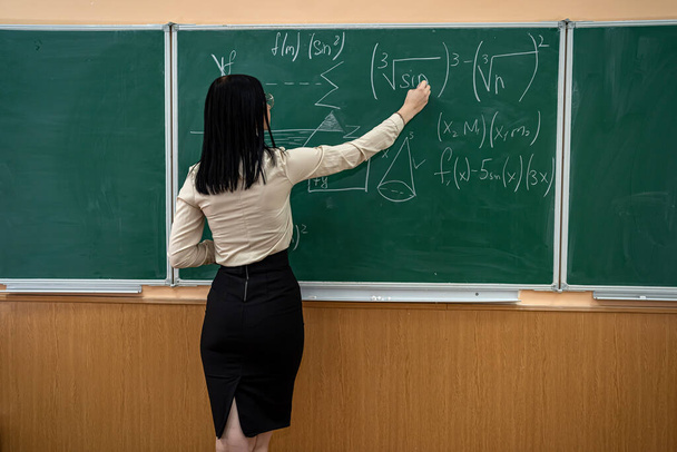 Hermosa maestra cerca de pizarra aprender matemáticas en el aula, vista trasera - Foto, imagen
