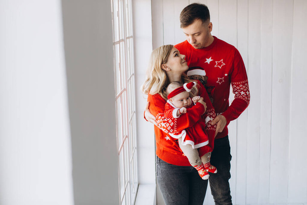 Happy family mom dad and little daughter in red traditional christmas outfit spending time together in light wooden room near window, copy space - Photo, Image