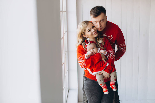 Feliz familia mamá papá y su hija pequeña en traje de Navidad tradicional rojo pasar tiempo juntos en la habitación de madera de luz cerca de la ventana, espacio de copia - Foto, Imagen