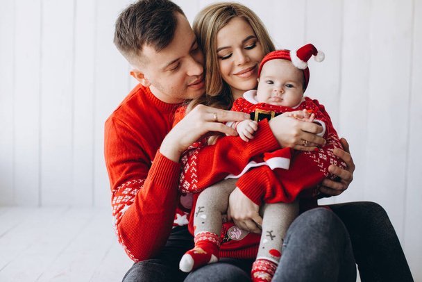 Portrait of happy family mom dad and little daughter in red traditional christmas clothes spending time together in light wooden room near window, copy space - Photo, Image