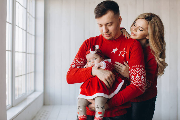 Портрет щасливої сім'ї мама тато і маленька дочка в червоному традиційному різдвяному одязі, що проводить час разом у світлій дерев'яній кімнаті біля вікна, копіювання простору
 - Фото, зображення