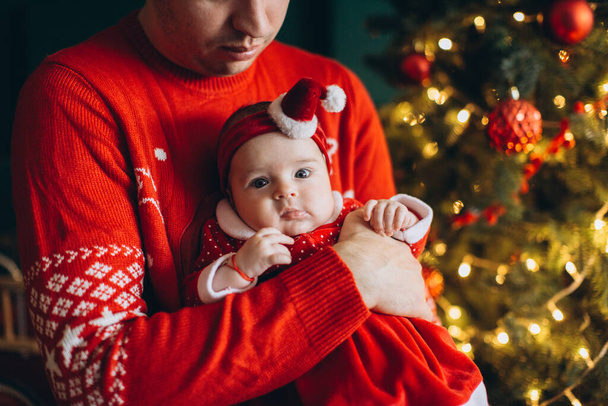 Fragment of a portrait of a dad and a little baby girl in traditional Christmas clothes near the Christmas tree - Foto, afbeelding